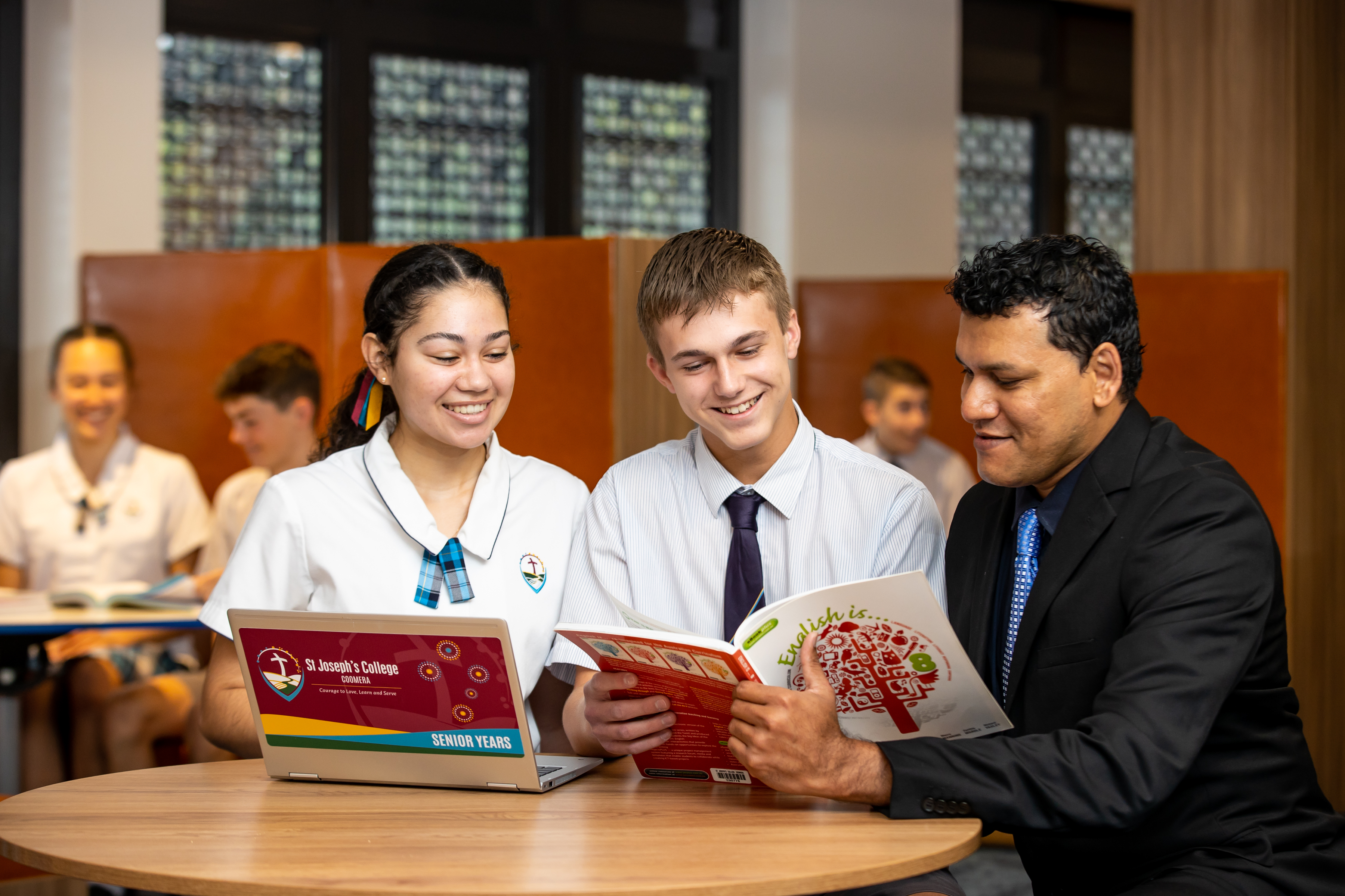 Two students and an adult seated at a table in a classroom, reading a book together with a laptop open beside them.